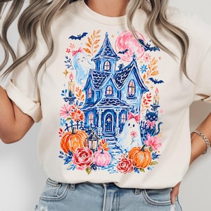 May include: A cream-colored t-shirt featuring a colorful graphic print of a blue haunted house, two ghosts with pink bows, a black cat, orange pumpkins, red and pink flowers, bats, and a pink moon. The design includes candles and a lantern.