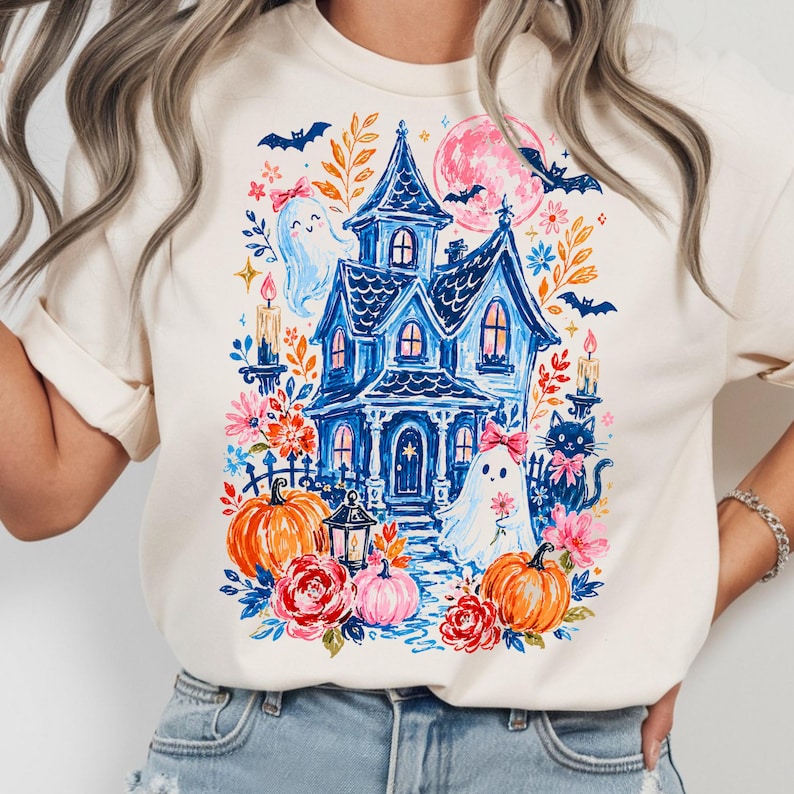 May include: A cream-colored t-shirt featuring a colorful graphic print of a blue haunted house, two ghosts with pink bows, a black cat, orange pumpkins, red and pink flowers, bats, and a pink moon. The design includes candles and a lantern.