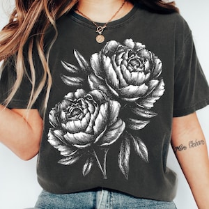 Puede incluir: Camiseta gris oscuro con una ilustración en blanco y negro de dos peonías y hojas. El diseño floral está centrado en la camiseta. La camiseta tiene cuello redondo y mangas cortas.