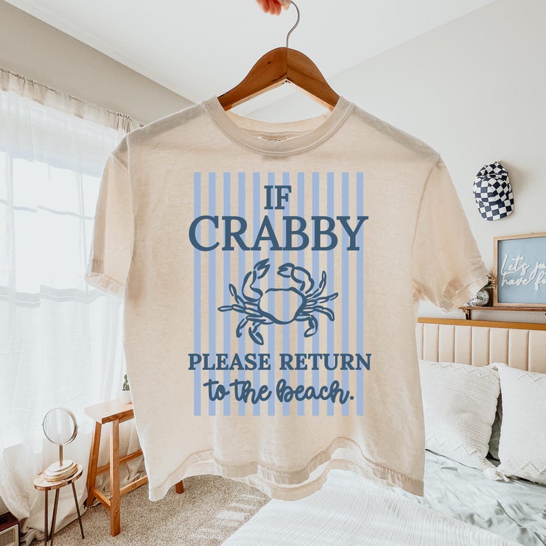 Puede incluir: Camiseta color crema con el texto "IF CRABBY PLEASE RETURN to the beach." El dise&ntilde;o presenta una ilustraci&oacute;n de cangrejo azul y rayas verticales azules. La camiseta est&aacute; colgada en una percha de madera.