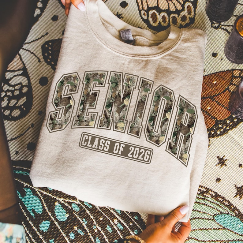Pu&ograve; includere: Felpa beige con la scritta "SENIOR" a grandi lettere a blocchi riempite con un motivo mimetico. Sotto c'&egrave; la scritta "CLASS OF 2026". La felpa &egrave; piegata e tenuta tra le mani.