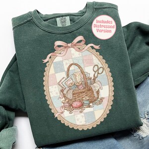 May include: A dark green sweatshirt with a detailed graphic of a sewing basket, scissors, spools of thread, and fabric scraps. The graphic is inside an oval frame with a pink bow. The text "Includes Distressed Version" is in a pink circle.