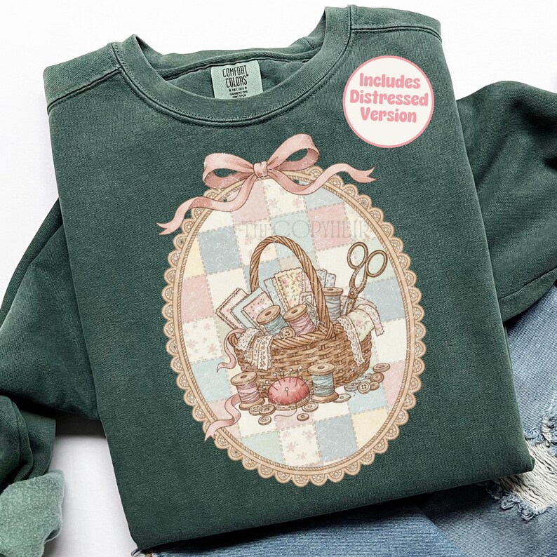 May include: A dark green sweatshirt with a detailed graphic of a sewing basket, scissors, spools of thread, and fabric scraps. The graphic is inside an oval frame with a pink bow. The text "Includes Distressed Version" is in a pink circle.