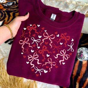 May include: Burgundy sweatshirt featuring a heart design made of red and white bows and hearts. The bows have polka dot and solid patterns. The sweatshirt is held by a person's hand.