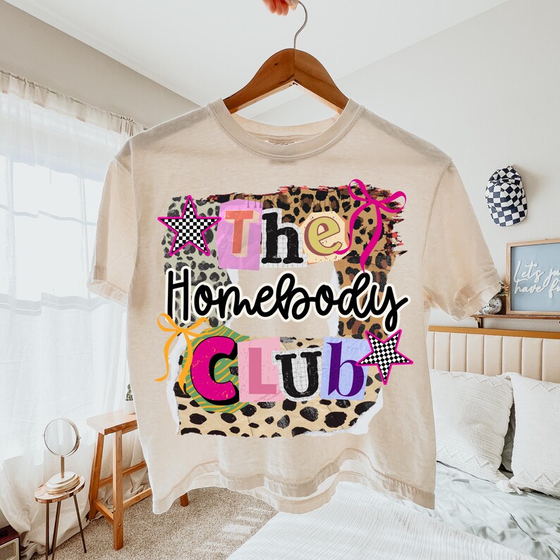 May include: A cream-colored t-shirt with the words "The Homebody Club" in a colorful, playful design. The graphic includes leopard print, stars, and bows. The shirt is hanging on a wooden hanger.