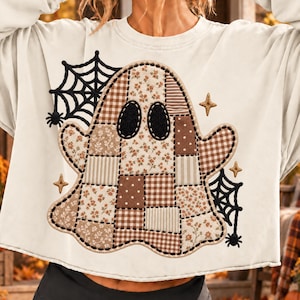 Può includere: Una felpa girocollo color crema con una grande grafica centrale raffigurante un fantasma. Il fantasma è realizzato con un motivo patchwork di tessuti marroni e bianchi a fiori, a quadretti e a pois. Ragnatele e stelle nere ricamate lo circondano.