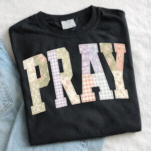 K&ouml;nnte beinhalten: Schwarzes T-Shirt mit dem Wort "PRAY" in Patchwork-Buchstaben. Die Buchstaben bestehen aus floralen und Vichy-Stoffen in Gr&uuml;n-, Lila-, Rosa- und Oranget&ouml;nen. Das T-Shirt ist auf einer wei&szlig;en Oberfl&auml;che gefaltet.