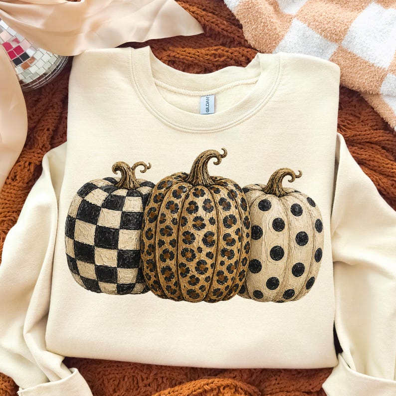 May include: Cream-colored sweatshirt featuring three decorative pumpkins. One pumpkin has a black and white checkerboard pattern, the second has a leopard print, and the third has black polka dots. The pumpkins have brown stems.