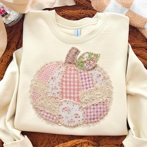 May include: A cream-coloured crewneck sweatshirt featuring a large, central pumpkin appliqué. The pumpkin is constructed from various patches of pink and white fabric, including gingham and floral patterns, accented with beige lace trim.