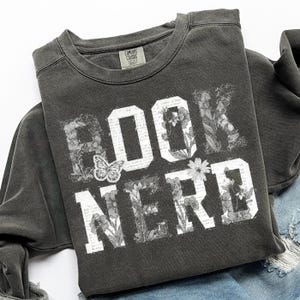 May include: Dark gray sweatshirt with the words "BOOK NERD" in white block letters. The letters are filled with floral and book page designs. A butterfly graphic is between the "O" and "K". The sweatshirt is folded on a light blue denim surface.