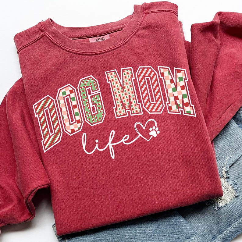 Może przedstawiać: Bordowa bluza z napisem "DOG MOM life" w ozdobnej czcionce. Litery zdobione są r&oacute;żnymi wzorami, w tym paskami i kratką. Serce i odcisk łapy towarzyszą słowu "life".