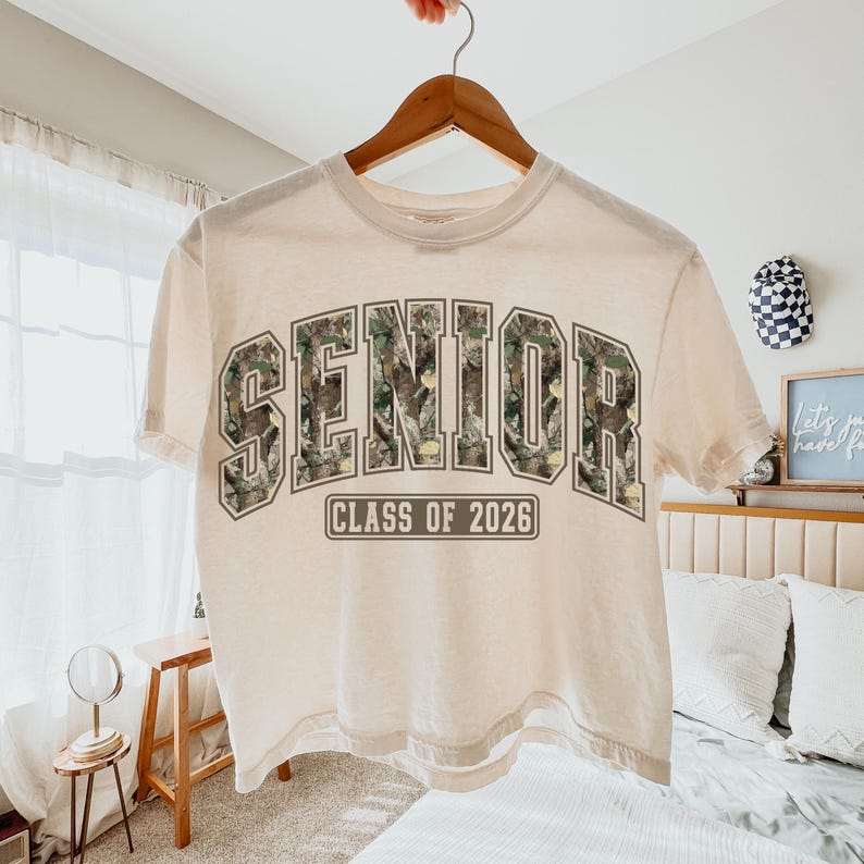 Pu&ograve; includere: Una maglietta color crema con la scritta "SENIOR" a grandi lettere con motivo mimetico. Sotto la parola "SENIOR" c'&egrave; il testo "CLASS OF 2026". La maglietta &egrave; appesa a una gruccia di legno.