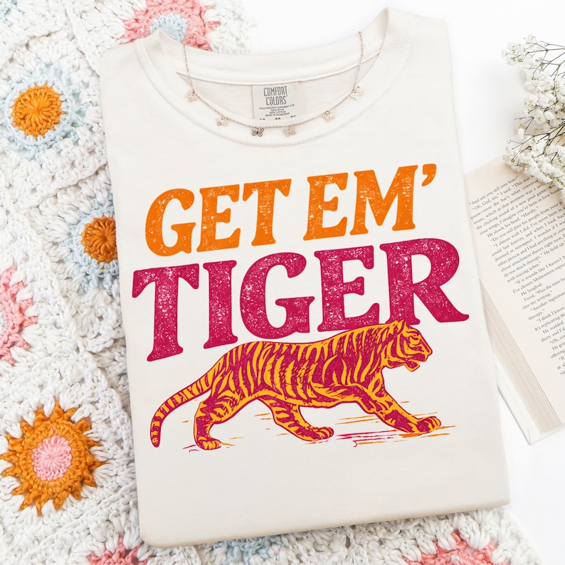 K&ouml;nnte beinhalten: Cremefarbenes T-Shirt mit dem Text "GET EM' TIGER" in Orange und Pink sowie einer Tigerillustration in Orange und Gelb. Eine silberne Halskette mit kleinen Anh&auml;ngern liegt auf dem Shirt.