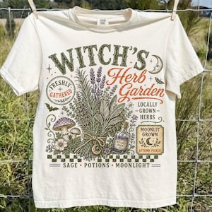 May include: A cream-colored short-sleeve t-shirt featuring a graphic print with the text Witch's Herb Garden. The design includes illustrations of bundled herbs, mushrooms, a potion bottle, a crescent moon, and a bat, with the words Sage, Potions, Moonlight.
