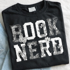 May include: Black t-shirt with the words "BOOK NERD" in white block letters. The letters are filled with floral and botanical illustrations, and a butterfly graphic is centered on the shirt.