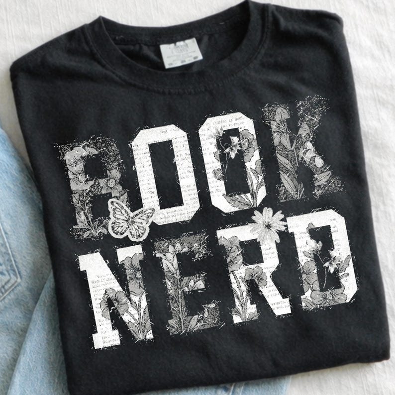 May include: Black t-shirt with the words "BOOK NERD" in white block letters. The letters are filled with floral and botanical illustrations, and a butterfly graphic is centered on the shirt.