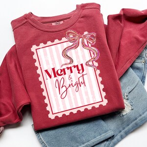 Puede incluir: Una sudadera granate con la frase "Merry & Bright" en letras rojas y rosas. El dise&ntilde;o es un dise&ntilde;o de sello postal con un fondo a rayas rosas y blancas y un lazo colorido. La sudadera est&aacute; doblada sobre unos vaqueros azules.