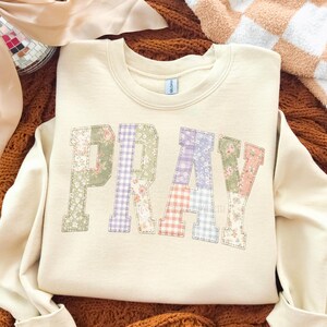 K&ouml;nnte beinhalten: Cremefarbenes Sweatshirt mit dem Wort "PRAY" in Patchwork-Buchstaben. Die Buchstaben bestehen aus verschiedenen Blumen- und Vichy-Mustern in Pastellfarben. Das Sweatshirt wird auf einer braunen Oberfl&auml;che pr&auml;sentiert.