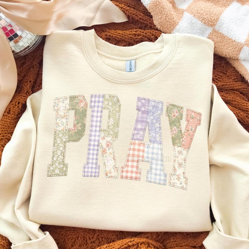 K&ouml;nnte beinhalten: Cremefarbenes Sweatshirt mit dem Wort "PRAY" in Patchwork-Buchstaben. Die Buchstaben bestehen aus verschiedenen Blumen- und Vichy-Mustern in Pastellfarben. Das Sweatshirt wird auf einer braunen Oberfl&auml;che pr&auml;sentiert.