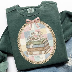 May include: Dark green sweatshirt with a vintage graphic. The graphic shows a teacup and saucer on a stack of books, with a pink bow and a decorative border. The text "THECOPYHEIR" is at the bottom. The sweatshirt has a crew neck and long sleeves.