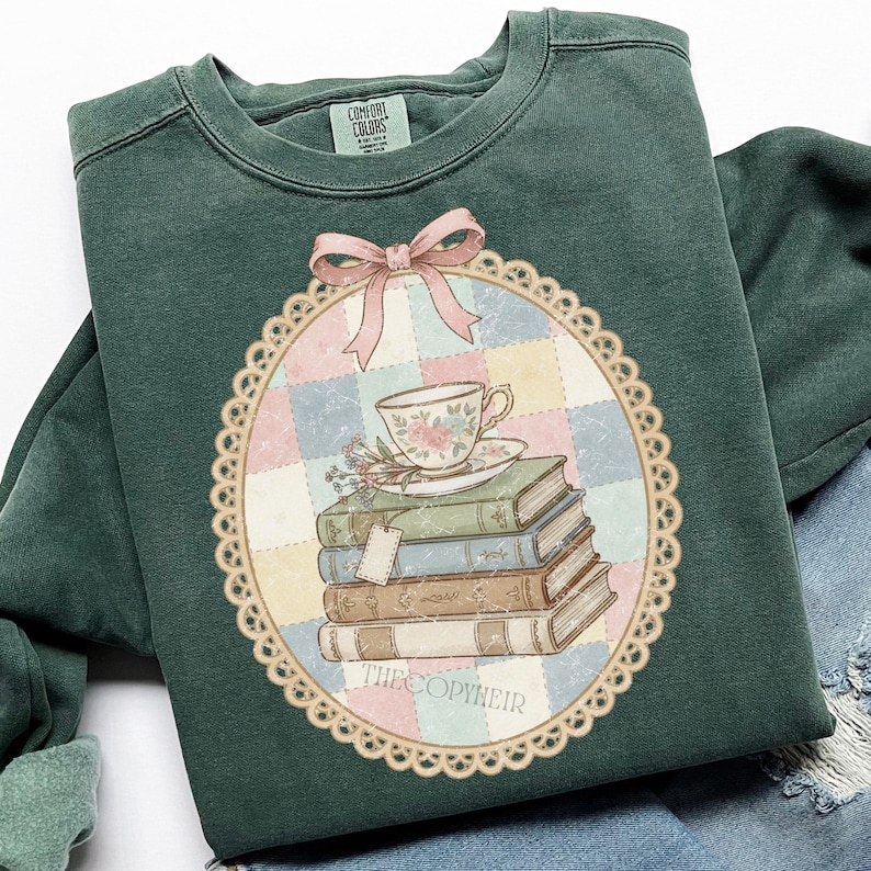 May include: Dark green sweatshirt with a vintage graphic. The graphic shows a teacup and saucer on a stack of books, with a pink bow and a decorative border. The text "THECOPYHEIR" is at the bottom. The sweatshirt has a crew neck and long sleeves.