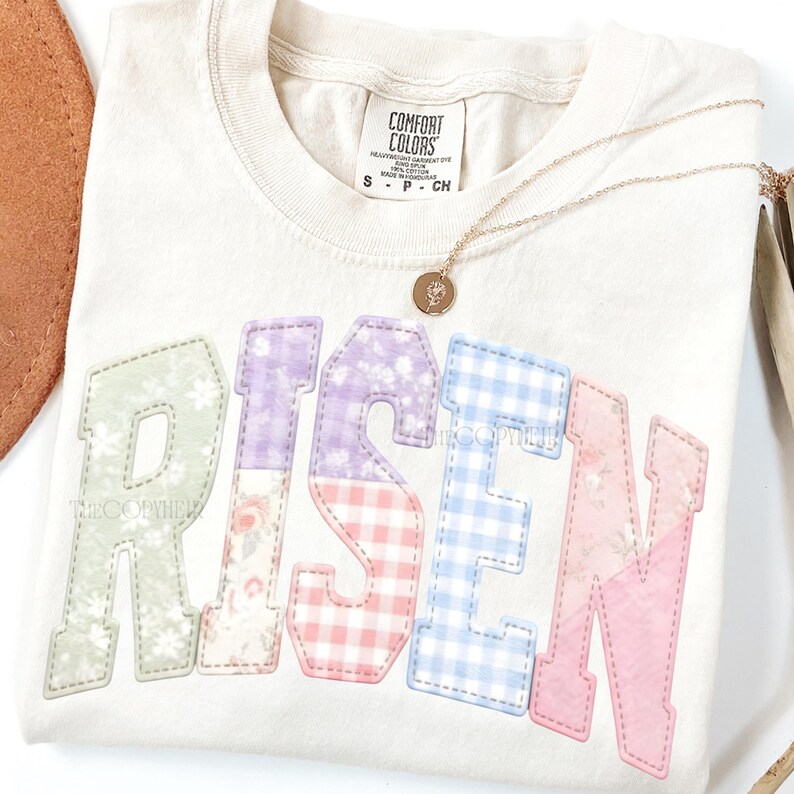 May include: Cream-colored Comfort Colors t-shirt with the word "RISEN" in pastel patchwork letters. A gold necklace with a pendant rests on the shirt. The letters are in green, purple, pink, and blue patterns.