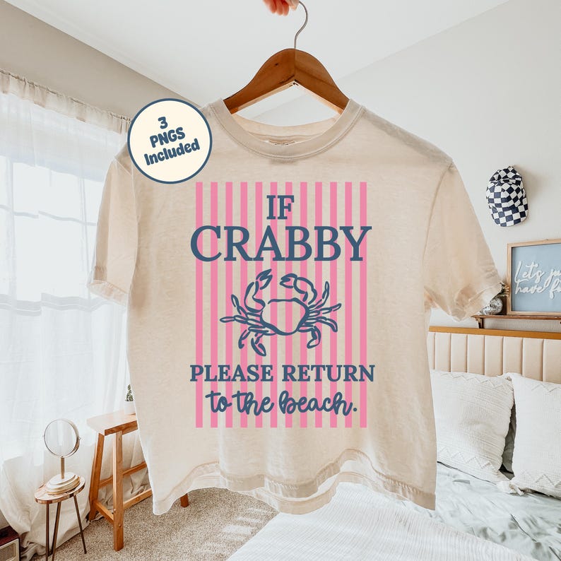 Puede incluir: Una camiseta color crema con el texto "IF CRABBY PLEASE RETURN to the beach" en azul y rosa. La camiseta presenta un gr&aacute;fico de cangrejo y est&aacute; colgada de una percha de madera. Un peque&ntilde;o c&iacute;rculo a la izquierda dice "3 PNGs Included".