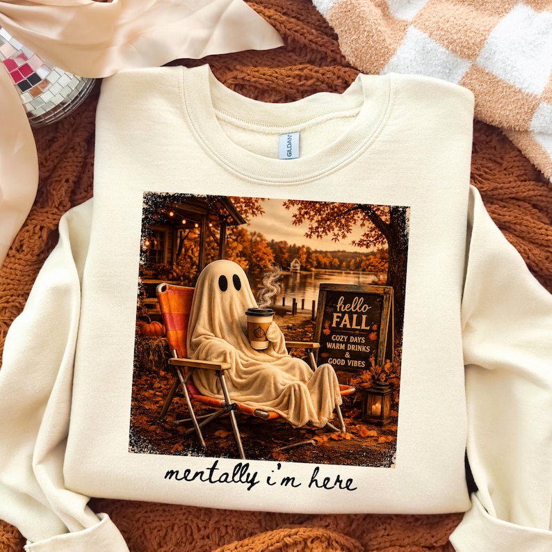 May include: Cream-colored sweatshirt featuring a graphic of a ghost in a sheet, holding a coffee cup, relaxing in a chair by a lake. The image includes the text "hello FALL" and "mentally i'm here".