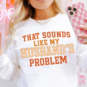 May include: White sweatshirt with the text "THAT SOUNDS LIKE MY HUSBAND'S PROBLEM" in orange and peach lettering. The sweatshirt is a crew neck style. The image also shows a gold chain necklace and a pink and white checkered phone case.
