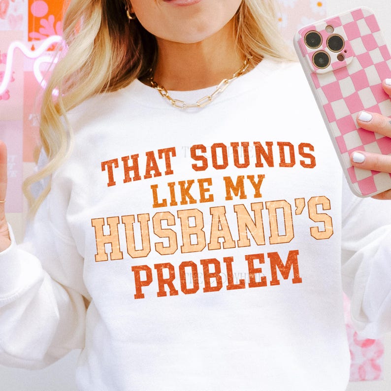 May include: White sweatshirt with the text "THAT SOUNDS LIKE MY HUSBAND'S PROBLEM" in orange and peach lettering. The sweatshirt is a crew neck style. The image also shows a gold chain necklace and a pink and white checkered phone case.