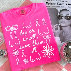 May include: A bright pink t-shirt with white text and illustrations. The text reads "big or small, save them all" with drawings of bows, flowers, and breasts. The shirt is on a pink and white checkered surface.