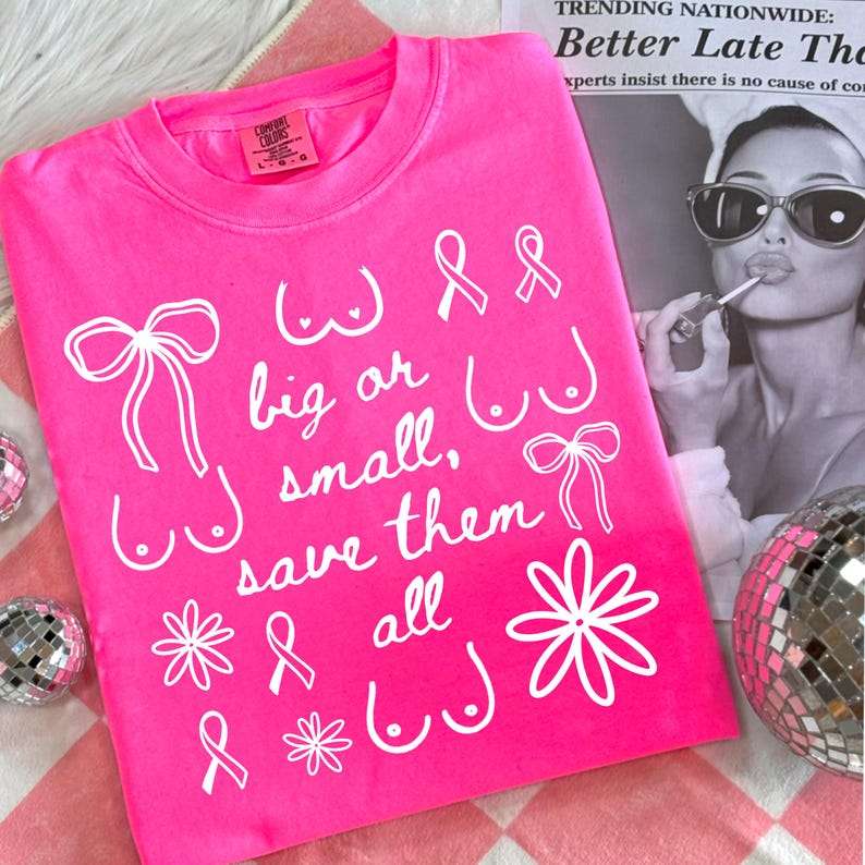 May include: A bright pink t-shirt with white text and illustrations. The text reads "big or small, save them all" with drawings of bows, flowers, and breasts. The shirt is on a pink and white checkered surface.