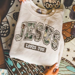 K&ouml;nnte beinhalten: Beiges Sweatshirt mit dem Wort "JESUS" in gro&szlig;en Buchstaben mit Tarnmuster. Darunter steht in einem braunen Rechteck "LOVES YOU". Das Sweatshirt ist gefaltet und wird von H&auml;nden gehalten.
