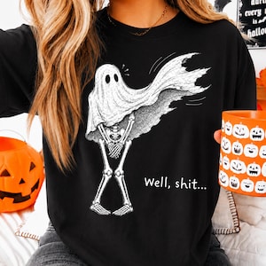 Op de afbeelding: Een zwart t-shirt met een witte grafische illustratie van een skelet in een spooklaken. Het skelet staat met gekruiste benen en houdt de handen bij het bekken. De tekst "Well, shit..." staat in het wit aan de rechterkant van het skelet.