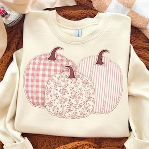 May include: A cream-colored crewneck sweatshirt featuring a graphic of three pumpkins. The left pumpkin has a pink and white gingham pattern, the center one has a pink floral print, and the right one has a pink and white vertical stripe pattern.