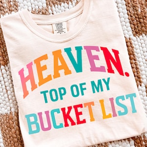 May include: Cream-colored t-shirt with the words "HEAVEN. TOP OF MY BUCKET LIST" in colorful, bold letters. The shirt is folded and displayed on a patterned surface. The shirt has a Comfort Colors tag.