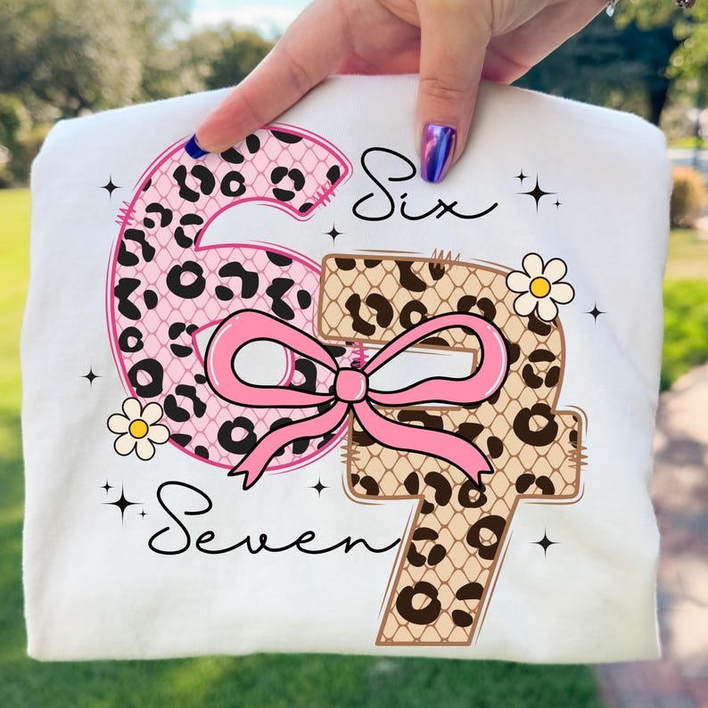 Pode incluir: Camiseta branca com os n&uacute;meros "6" e "7" em rosa e estampa de leopardo. Um la&ccedil;o rosa une os n&uacute;meros, com as palavras "Six" e "Seven" em it&aacute;lico. O design tamb&eacute;m inclui pequenas flores e estrelas.