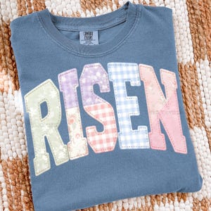 May include: A blue Comfort Colors crew neck t-shirt with the word "RISEN" in a patchwork design. The letters are in various patterns, including floral, gingham, and solid colors. The shirt is folded on a woven surface.