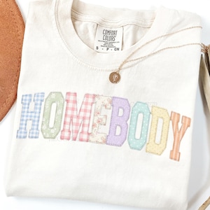 May include: Off-white t-shirt featuring the word "HOMEBODY" in patchwork lettering. A gold necklace with a pendant is draped across the shirt. The shirt has a Comfort Colors label.