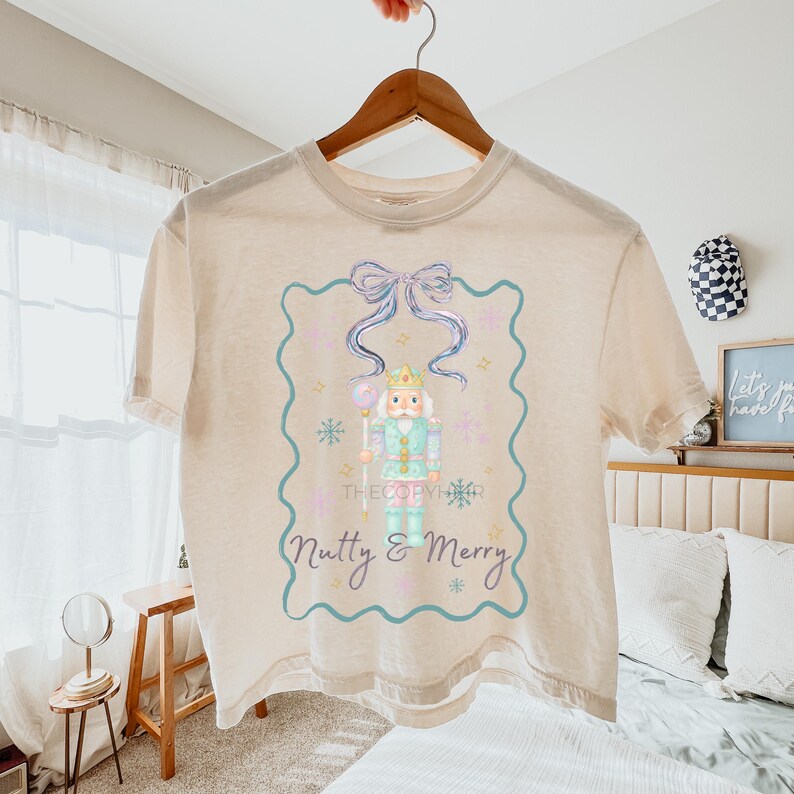 K&ouml;nnte beinhalten: Cremefarbenes T-Shirt mit einer Aquarellillustration eines Nussknackersoldaten. Das Design umfasst eine blaue Schleife, Schneeflocken und den Text "Nutty & Merry". Das T-Shirt h&auml;ngt an einem Holzb&uuml;gel.