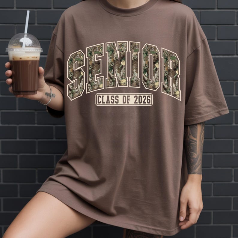 Pu&ograve; includere: T-shirt marrone con la scritta "SENIOR" in un motivo mimetico, bordata di bianco, sopra "CLASS OF 2026" in un rettangolo bianco. La maglietta &egrave; oversize e ha maniche corte. Una persona tiene in mano una bevanda con una cannuccia.