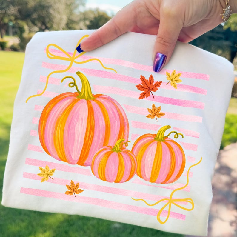 May include: White fabric with a fall-themed design. The design features three pumpkins in shades of pink and orange, with yellow stems and accents. Pink horizontal stripes and autumn leaves complete the seasonal artwork.