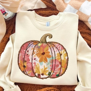 Peut inclure: Sweat-shirt crème avec un motif de citrouille florale. La citrouille est remplie de fleurs roses, oranges et jaunes. Le sweat-shirt a un col rond et des manches longues, parfait pour l'automne.