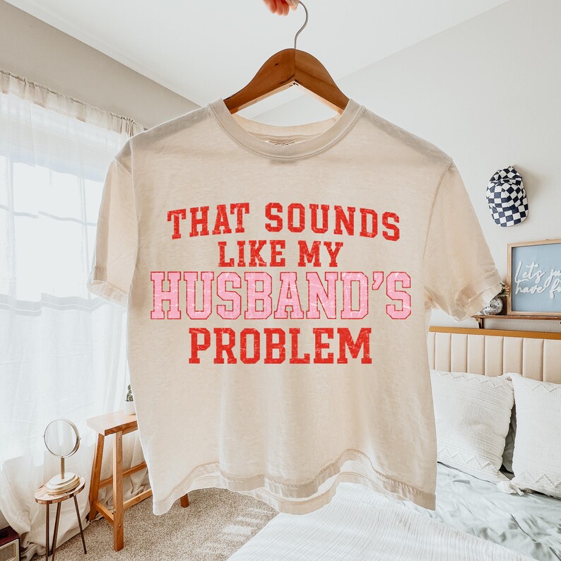 May include: Cream-colored t-shirt with the phrase "THAT SOUNDS LIKE MY HUSBAND'S PROBLEM" in red and pink block letters. The shirt is hanging on a wooden hanger. The background includes a bed and a small side table.