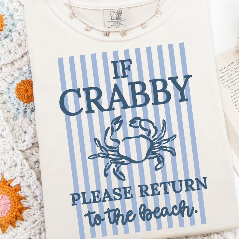 Puede incluir: Camiseta color crema con fondo a rayas azules y el texto "If Crabby Please Return to the beach." Un gr&aacute;fico de cangrejo azul est&aacute; centrado en la camiseta. Un delicado collar con peque&ntilde;os dijes est&aacute; en la parte superior.