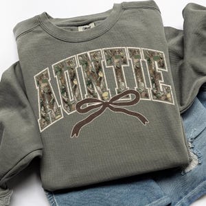 May include: An olive green sweatshirt with the word "AUNTIE" in white block letters filled with a camouflage pattern. A brown bow graphic is below the text. The sweatshirt is folded on top of a pair of denim shorts.