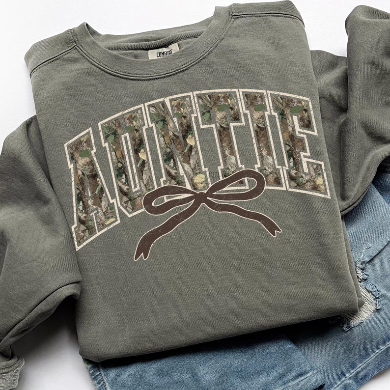 May include: An olive green sweatshirt with the word "AUNTIE" in white block letters filled with a camouflage pattern. A brown bow graphic is below the text. The sweatshirt is folded on top of a pair of denim shorts.