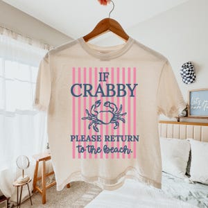 Puede incluir: Una camiseta color crema con el texto "IF CRABBY PLEASE RETURN to the beach." El texto es en azul marino y rosa, con un gr&aacute;fico de cangrejo. La camiseta est&aacute; colgada en una percha de madera.