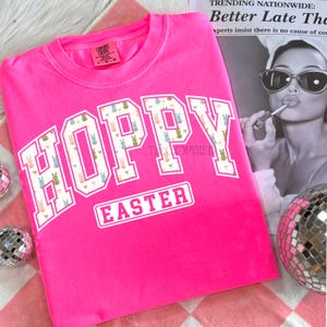 May include: A bright pink sweatshirt with the words "HOPPY EASTER" in white block letters. The letters "HOPPY" are filled with a pattern of pastel-colored Easter bunnies. The sweatshirt is a Comfort Colors brand.