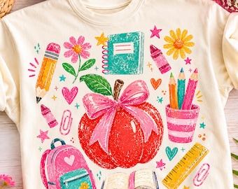 May include: A cream-colored t-shirt featuring a colorful school-themed graphic print. The design includes a large red apple with a pink bow, a pink backpack, a yellow pencil, a ruler, a notebook, colored pencils in a cup, and various flowers.